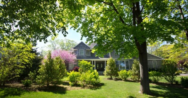 A suburban home with a manicured lawn, leafy trees, and flowering shrubs under bright sunlight in a quiet neighborhood.